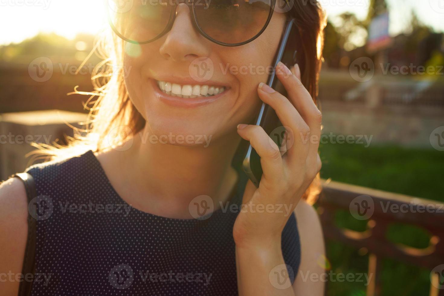 kvinna i solglasögon talande på de smartphone medan gående ner de gata på solnedgång, stänga upp foto