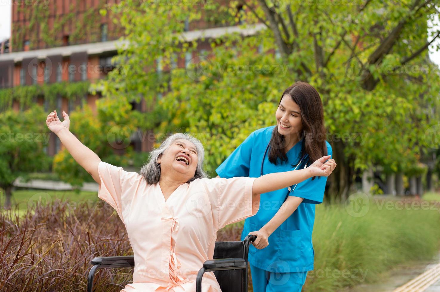 asiatisk försiktig vårdgivare eller sjuksköterska och de Lycklig patient i en rullstol är gående i de trädgård. begrepp av Lycklig pensionering med vård från en vårdgivare och besparingar och senior hälsa försäkring. foto