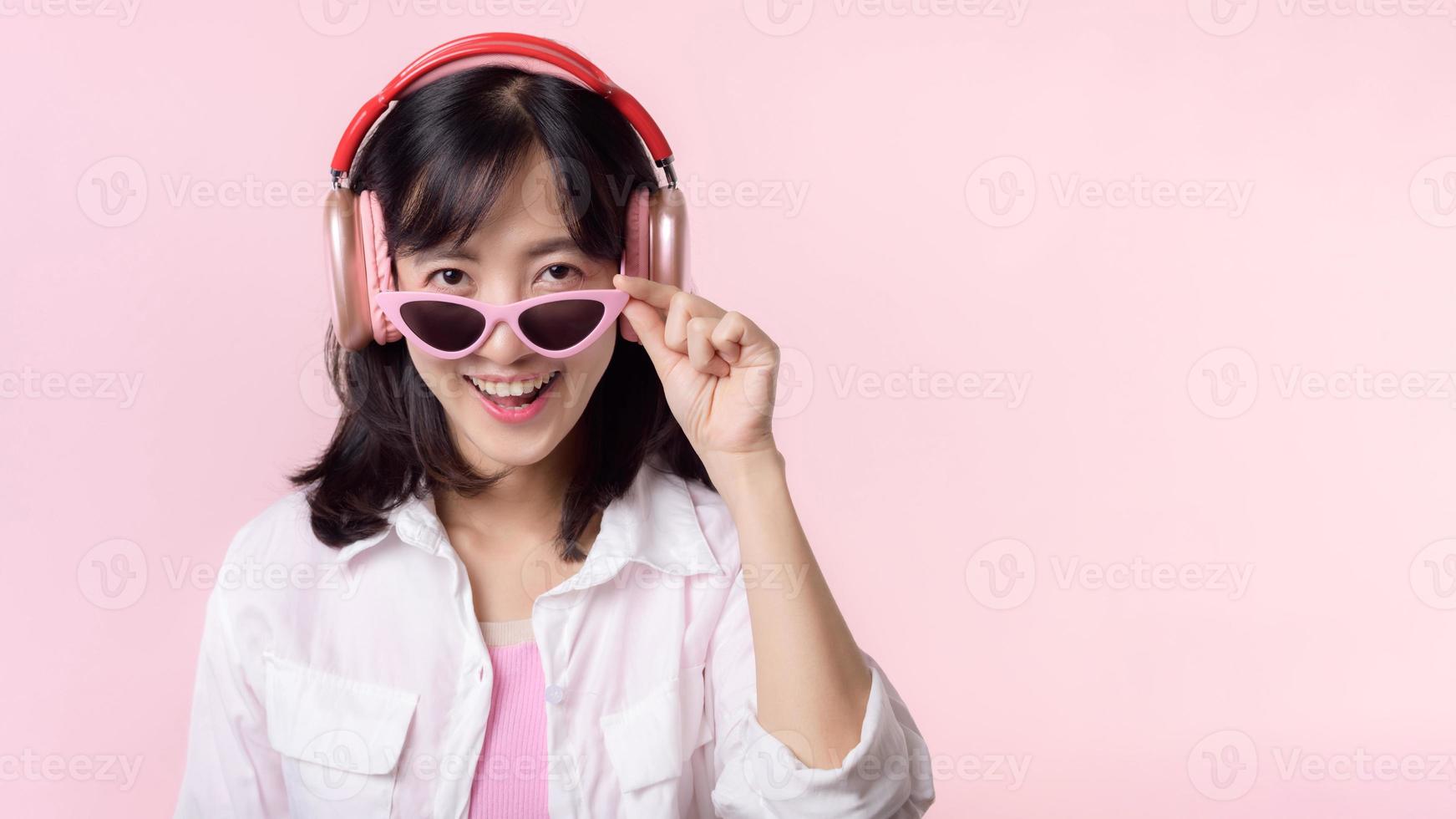Lycklig ung asiatisk kvinna modell med eleganta trendig Sol glasögon njut av lyssnande musik förbi hörlurar audio och dans isolerat på rosa studio bakgrund. teknologi, flicka mode, tillbehör begrepp. foto