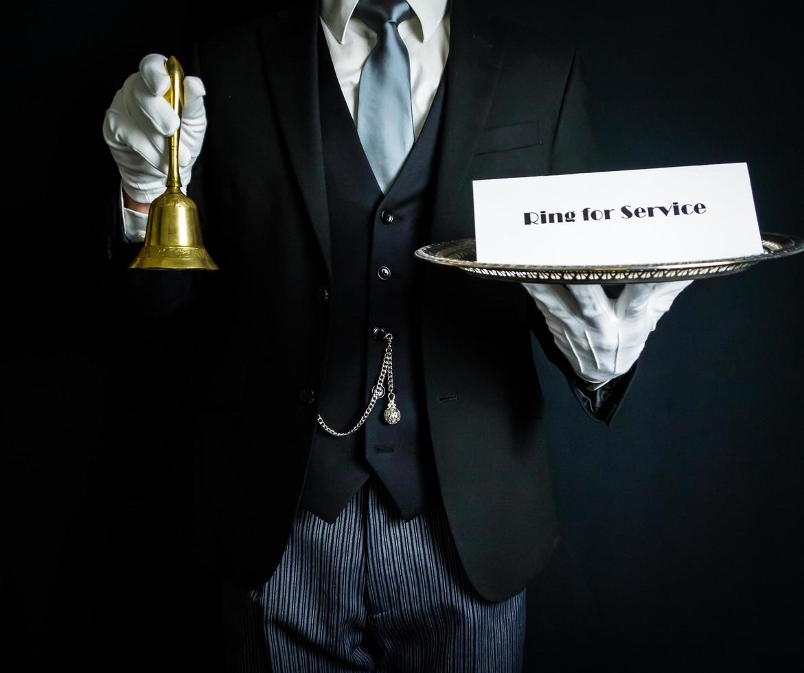 porträtt av butler i mörk formell kostym och vit handskar innehav klocka och tecken. ringa för service. begrepp av professionell gästfrihet. foto