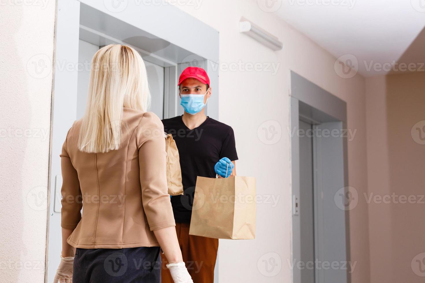 ung kvinna bär medicinsk mask tar emot beställd mat från de restaurang från leverans man inomhus. förebyggande av virus spridning foto