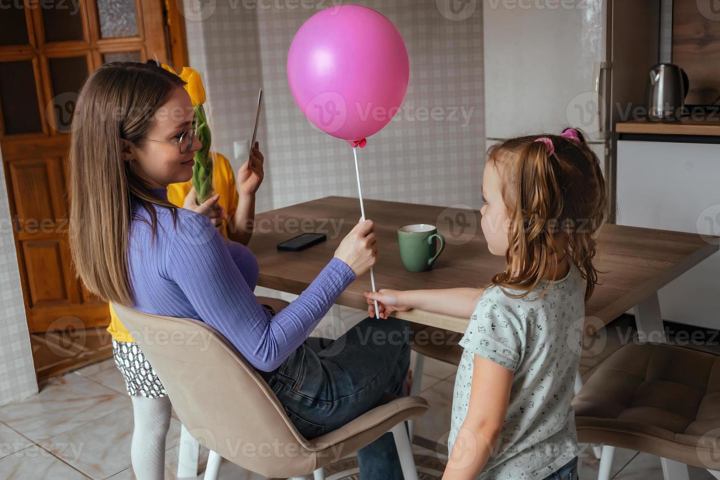 döttrar gratulera deras mamma på mors dag, en kort med en hjärta, blommor och en ballong på Hem i de kök. barn överraskning deras mor för de Semester. foto