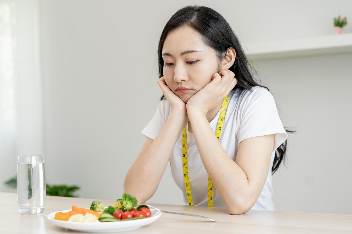 diet i uttråkad ansikte, olycklig skön asiatisk ung kvinna på bantning, ser på sallad tallrik på tabell, motvilja eller trött med äta färsk grönsaker. dietist av friska, näring av vikt förlust. foto