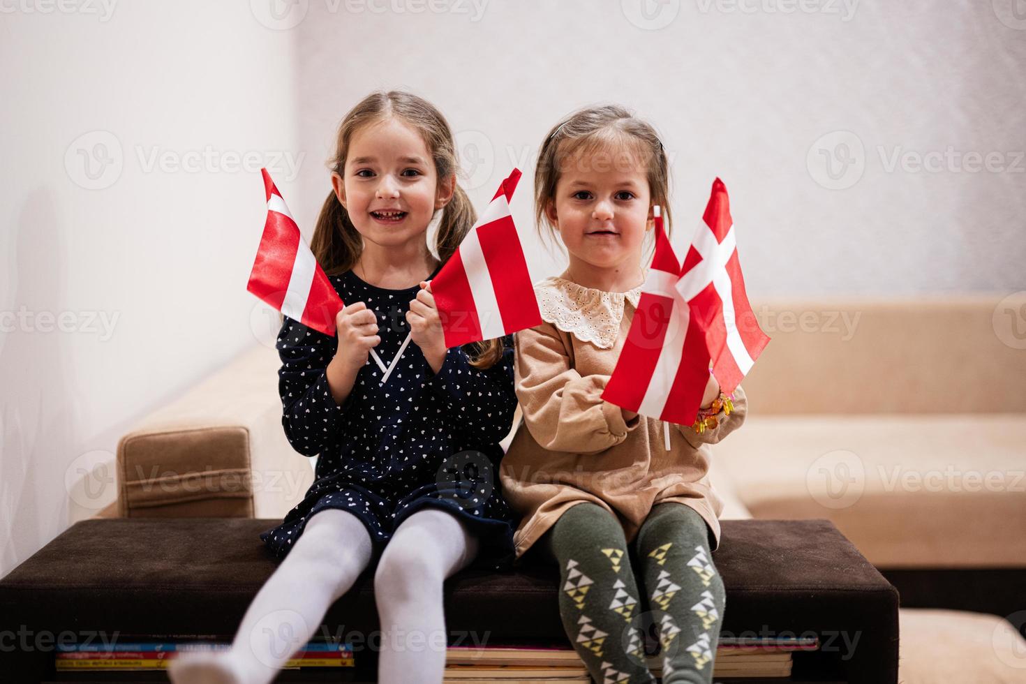 två systrar är Sammanträde på en soffa på Hem med dansk flaggor på händer. Danmark barn flickor med flagga . foto