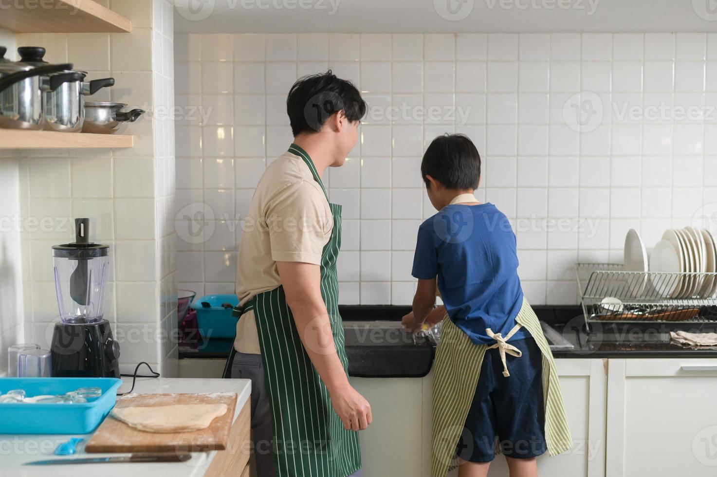 Lycklig leende ung asiatisk far och son tvättning maträtter i kök på Hem foto