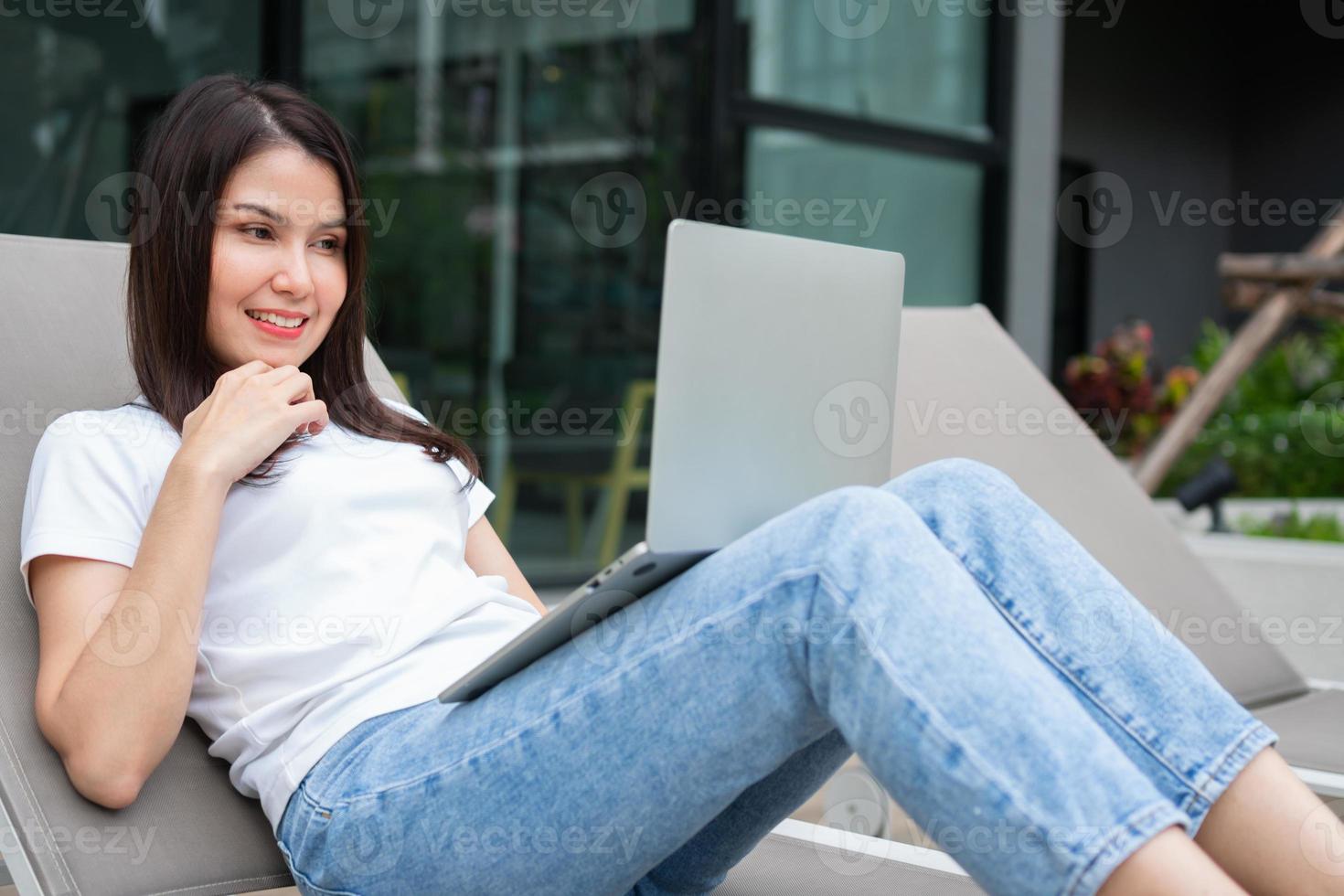 glad ung entreprenörskvinna som sitter på solariet bredvid poolen och använder bärbar dator för fjärranslutning online som arbetar digitalt, online affärsprojekt i en lugn gård till resorthuset, arbeta på semester foto