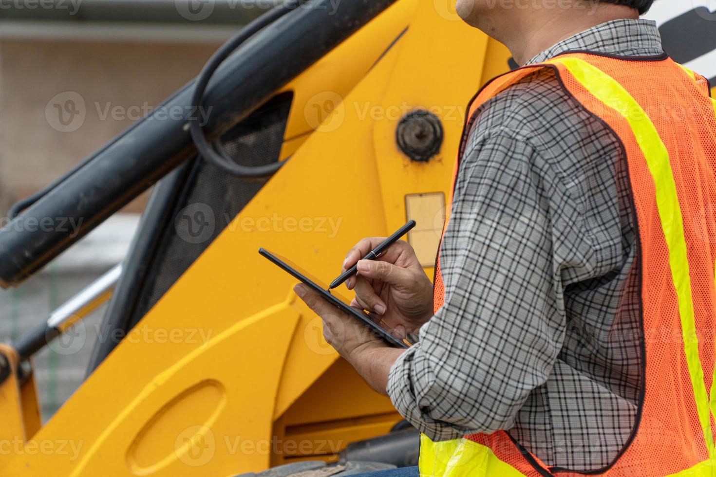 teknik bär en vit säkerhet hjälm stående i främre av de traktor ser på Hem konstruktion arbete och använda sig av de läsplatta till kolla upp de plan med konstruktion foto