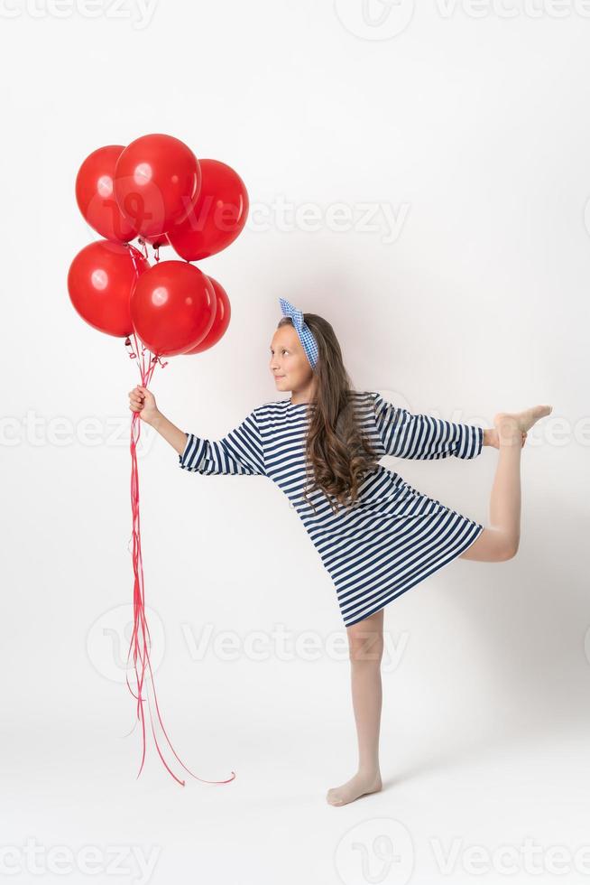 lekfull flicka innehav stor knippa av röd ballonger i hand, stående på ett ben, ser på ballonger foto