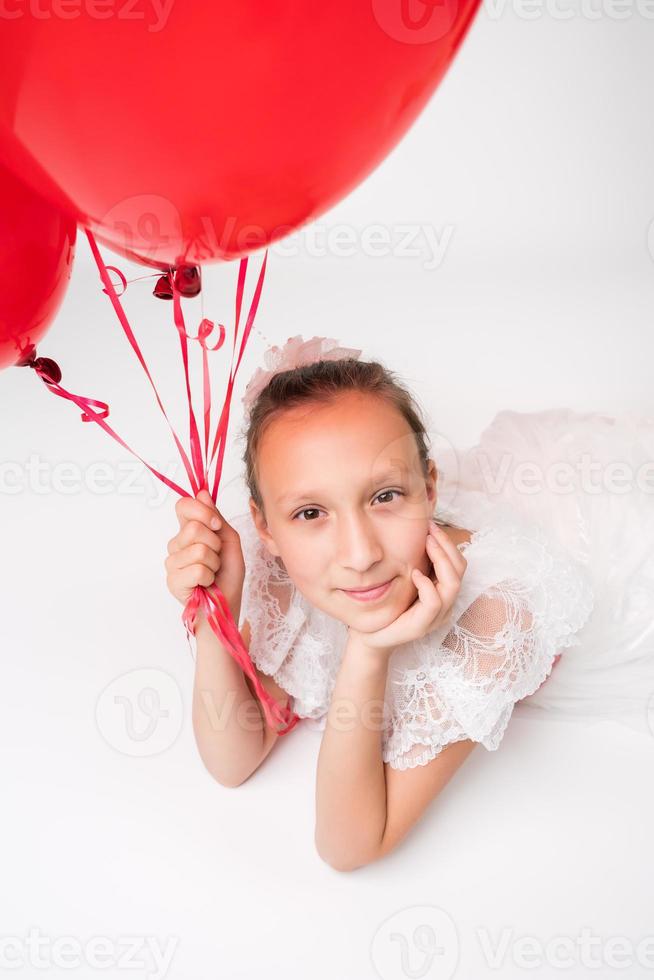 flicka innehav röd ballonger i hand och positiv ser på kamera, liggande ner på vit bakgrund foto