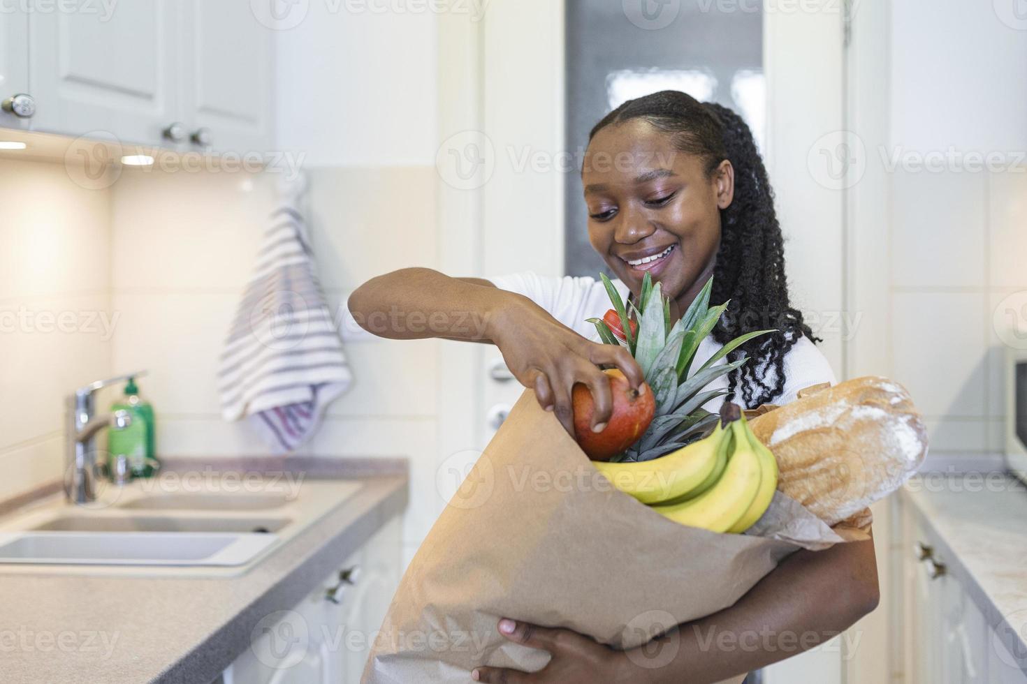 friska positiv Lycklig kvinna innehav en papper handla väska full av frukt och grönsaker. ung kvinna innehav matvaror handla väska med grönsaker stående i de kök foto