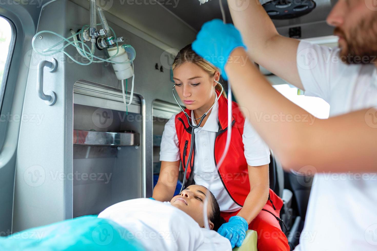 mediciner med skadade patient ger ett infusion . ung manlig paramedics innehav iv lösning. kvinna läkare listning hjärta av patient foto