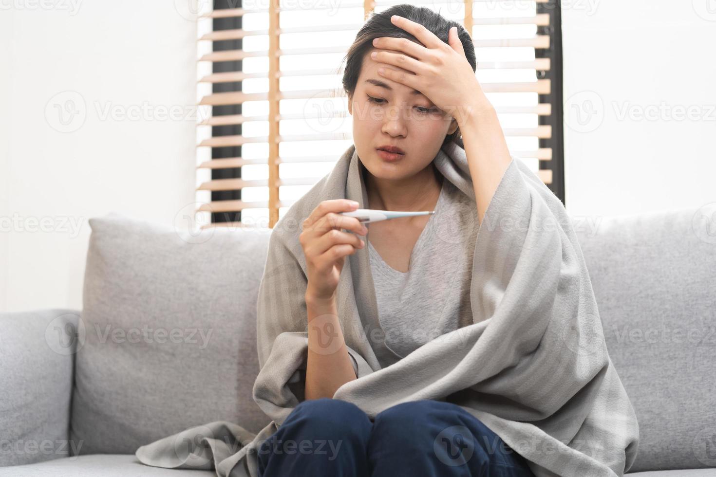 sjuk, influensa asiatisk ung kvinna, flicka huvudvärk ha en feber, influensa och kolla upp termometer mäta kropp temperatur, känna sjukdom Sammanträde på soffa säng på Hem. hälsa vård person på virus, covid19. foto