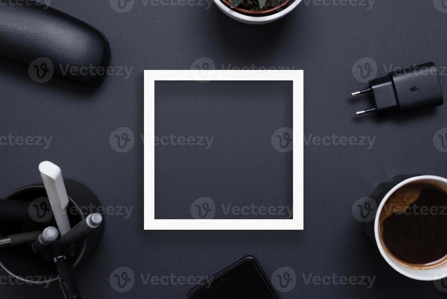 vit fyrkant papper ram på svart kontor skrivbord omgiven förbi kaffe mugg, laddare, växt, pennor och telefon. kopia Plats i de mitten. topp se platt lägga foto