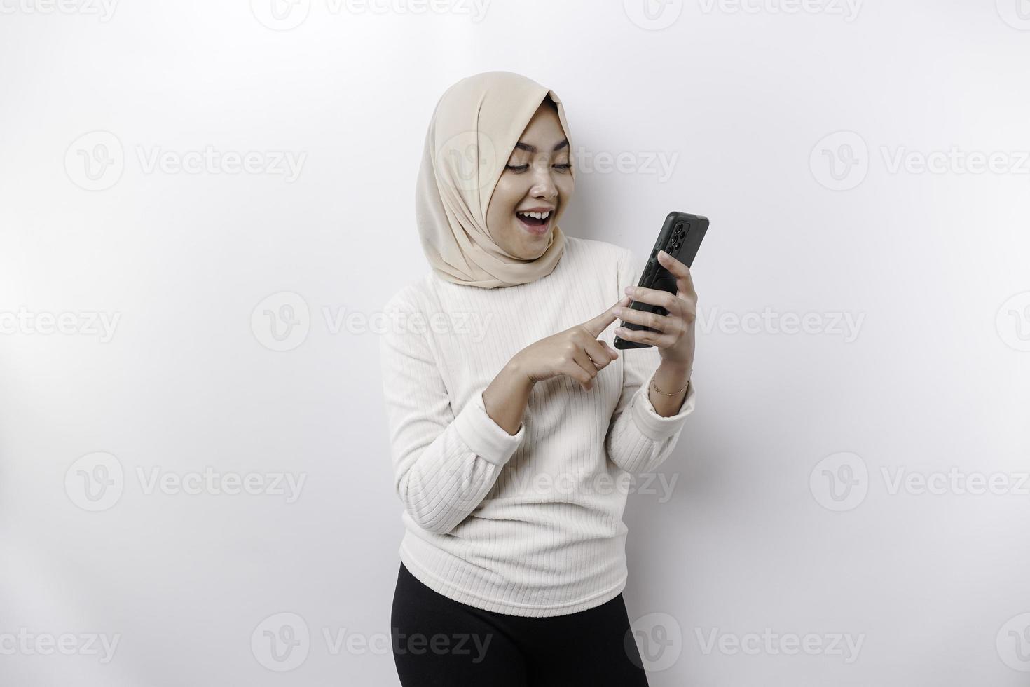 en Lycklig asiatisk muslim kvinna bär en huvudduk, innehav henne telefon, isolerat förbi vit bakgrund foto