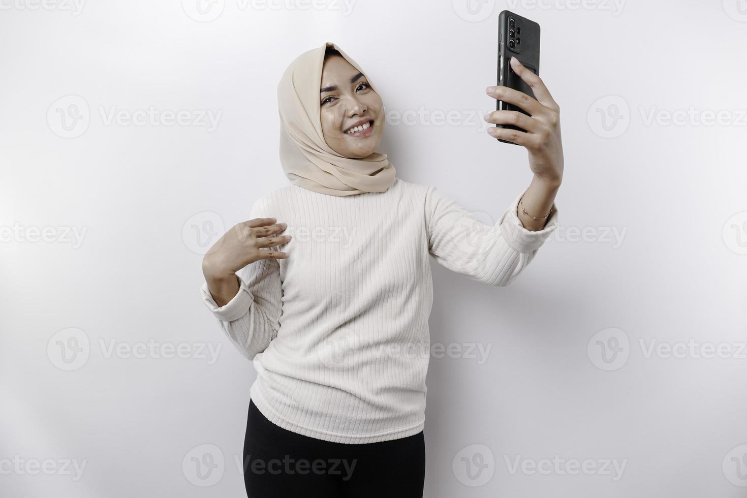 en Lycklig asiatisk muslim kvinna bär en huvudduk, innehav henne telefon, isolerat förbi vit bakgrund foto