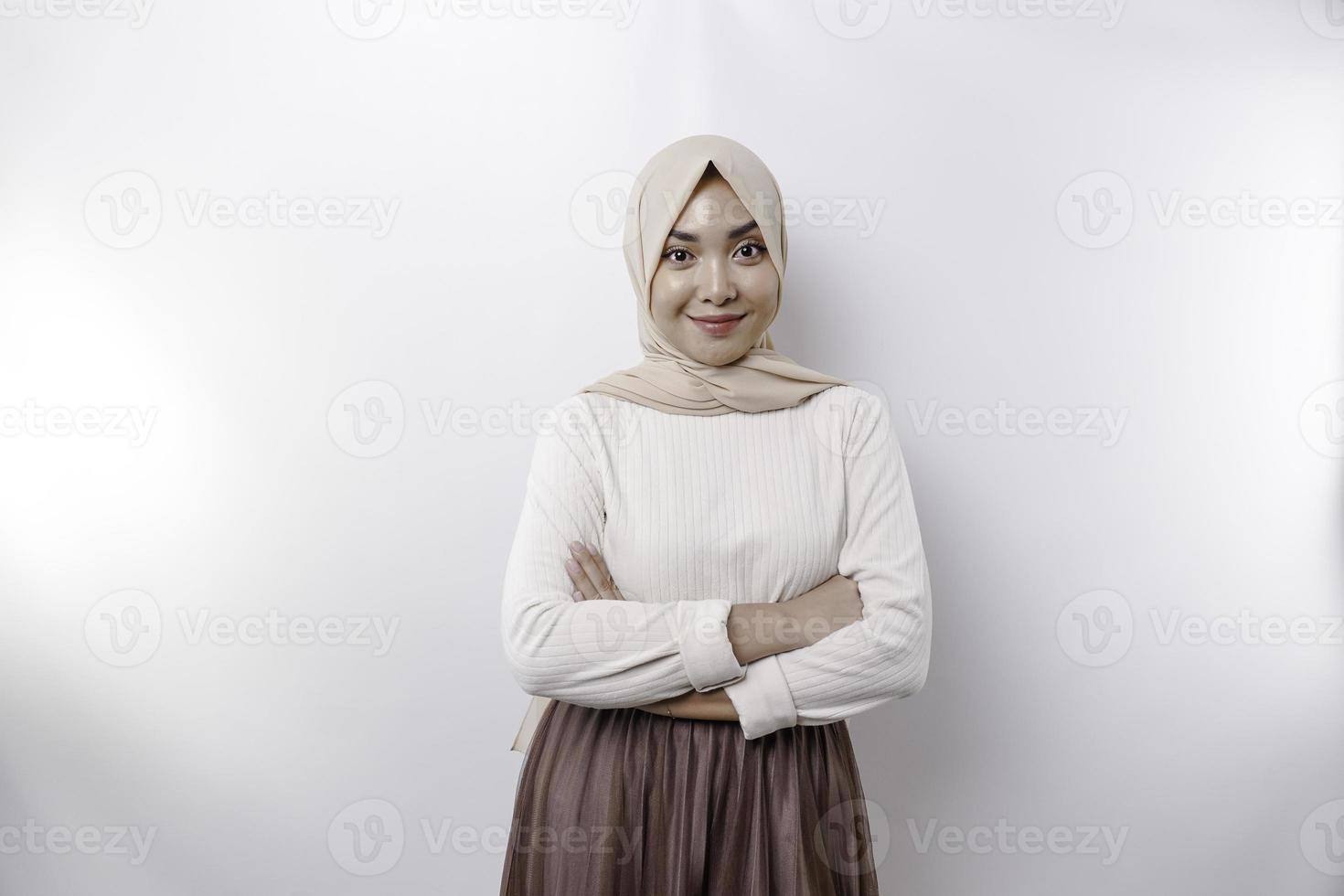 en självsäker leende asiatisk muslim kvinna bär hijab stående med vapen vikta och ser på de kamera isolerat över vit bakgrund foto