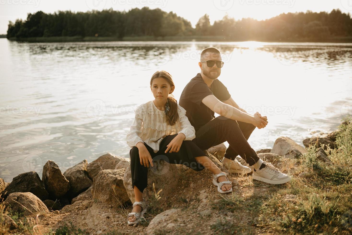 en ung far med skägg och solglasögon poserar på stenar med sin dotter vid kusten foto