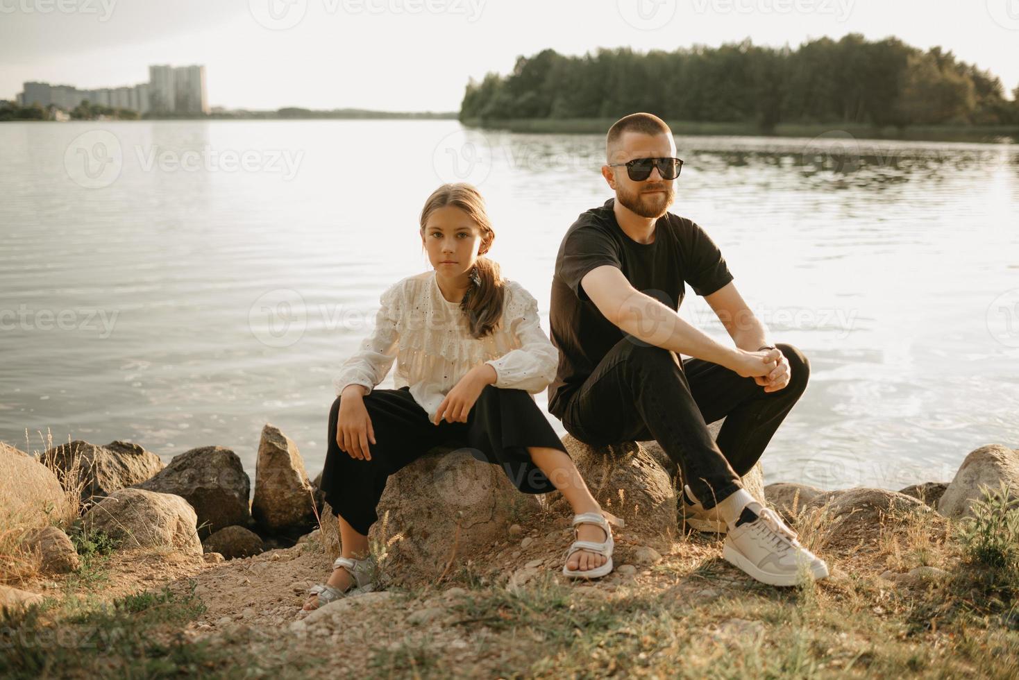 en ung far med skägg och solglasögon sitter på stenar med sin dotter vid kusten foto