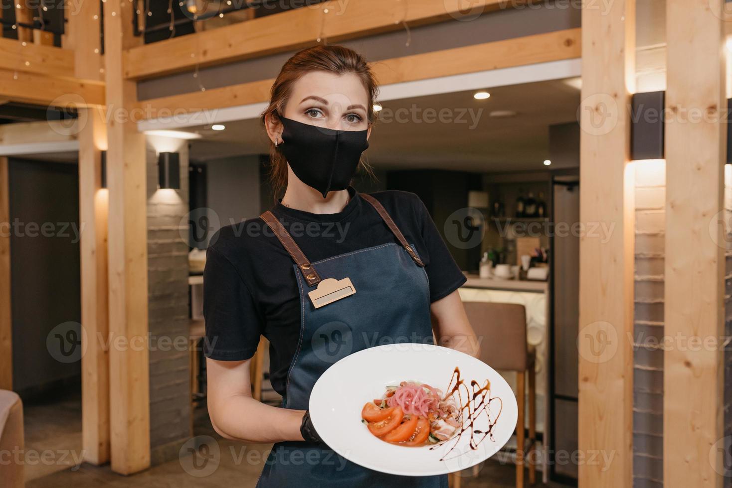 en snygg servitris som bär en svart ansiktsmask och engångshandskar håller en mynta och dolendwitz sallad på en restaurang foto