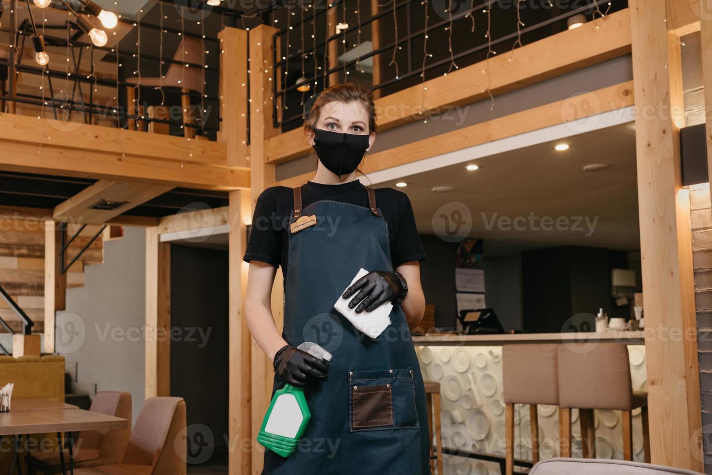 en servitris bär en medicinsk ansiktsmask och svarta medicinska engångshandskar som håller en flaska med desinfektionsmedel och en trasa för att rengöra bord foto