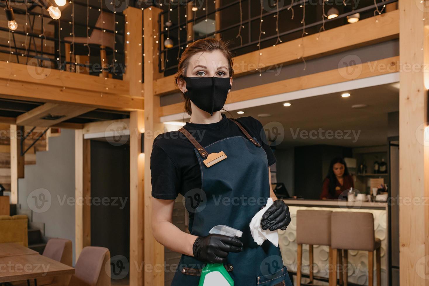 en snäll servitris bär en svart medicinsk ansiktsmask och medicinska handskar för engångsbruk som håller en flaska med desinfektionsmedel och rengöringsbord foto