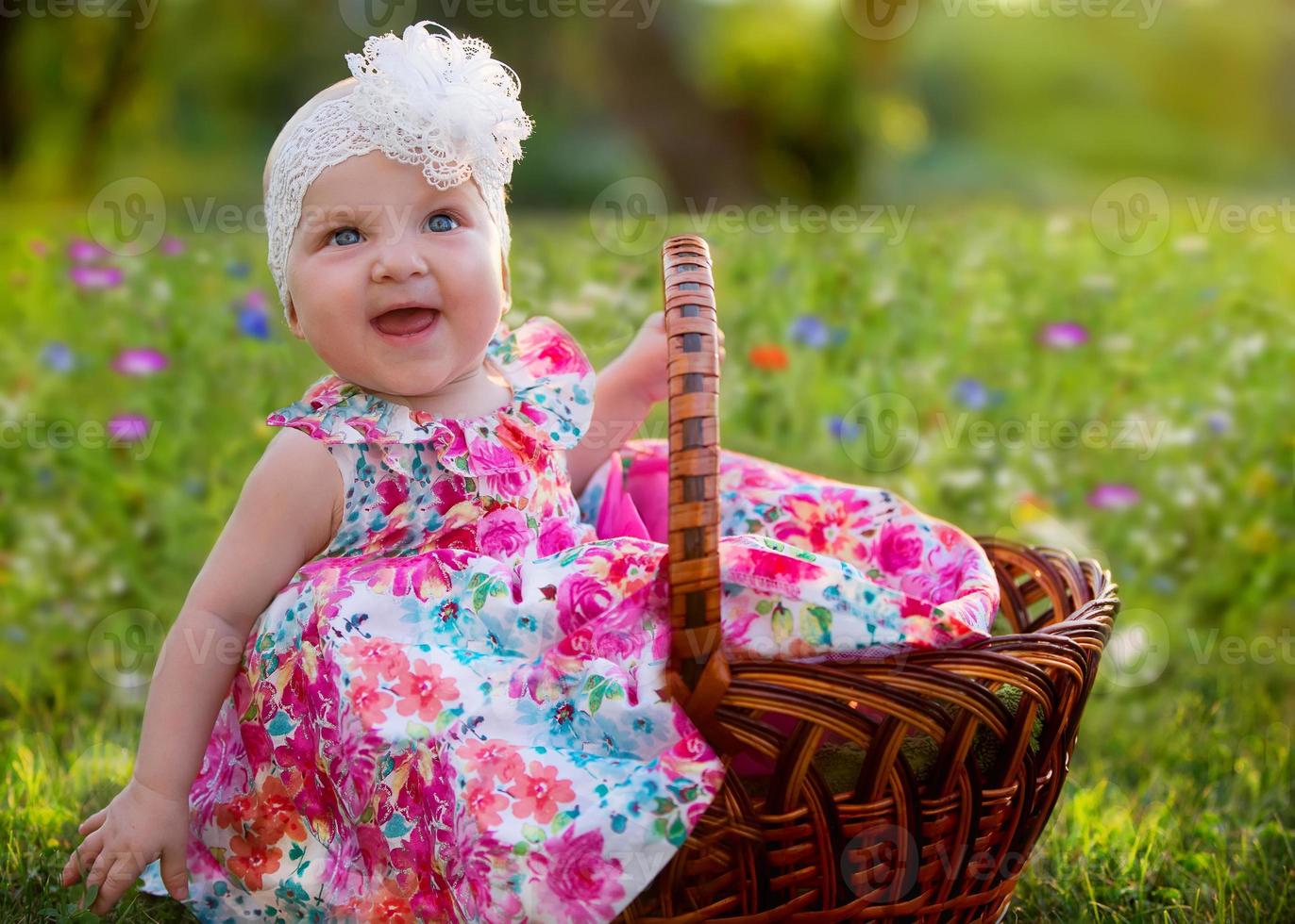en skön liten flicka sitter i en korg- korg och skrattar glatt på en blomma äng. skön barn. sommar promenad av en söt bebis. Lycklig barn upp till en år. foto