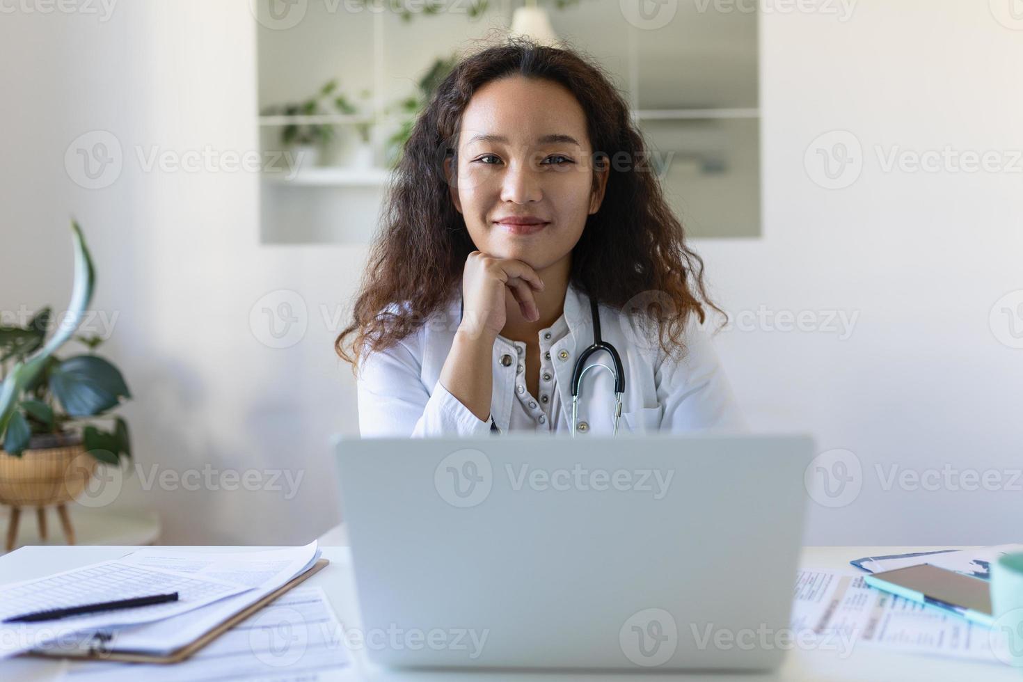 läkare professionell kvinna asiatisk läkare bär enhetlig tar anteckningar i medicinsk tidning, fyllning dokument, patient sjukdom historia, ser på bärbar dator skärm, studerande tittar på webinar foto