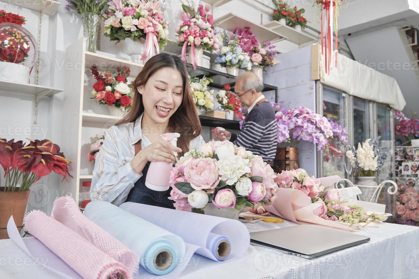 ung skön asiatisk kvinna blomsterhandlare arbetstagare vatten besprutning en flora knippa med gammal manlig anställd, Lycklig arbete i färgrik blomma affär Lagra, färsk blomma buketter dekorera, sme företag entreprenör. foto