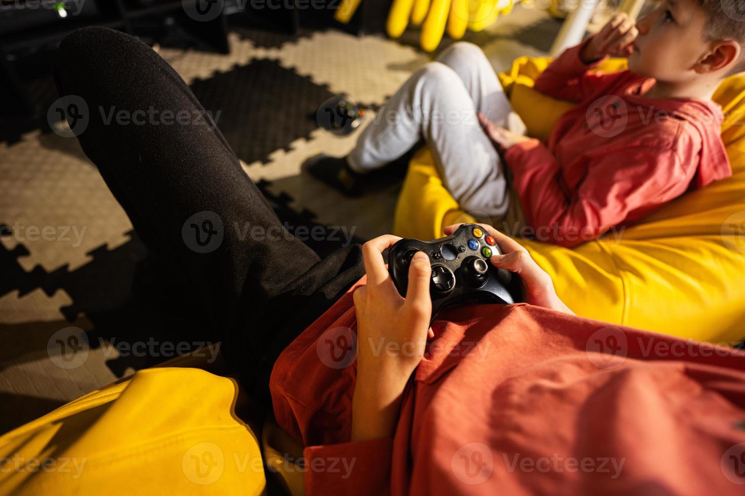 två bröder spelar video spel trösta, Sammanträde på gul pouf i barn spela Centrum. foto