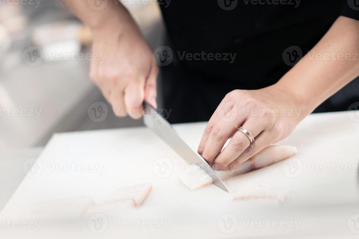 fokuserade manlig kock i enhetlig skärande fisk filea på styrelse, kock nedskärningar vit tilapia fisk i kök. foto