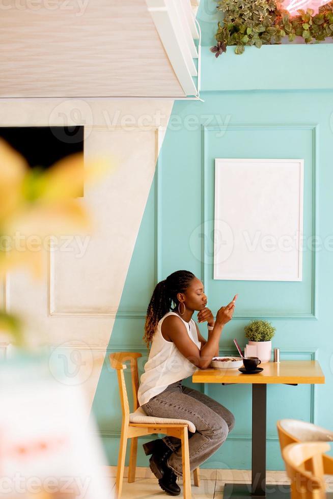 ung svart kvinna använder sig av mobil telefon medan har frukost i de Kafé foto