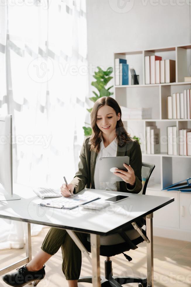 självsäker företag expert- attraktiv leende ung kvinna skriver bärbar dator ang innehav digital läsplatta på skrivbord i kreativ Hem kontor. foto