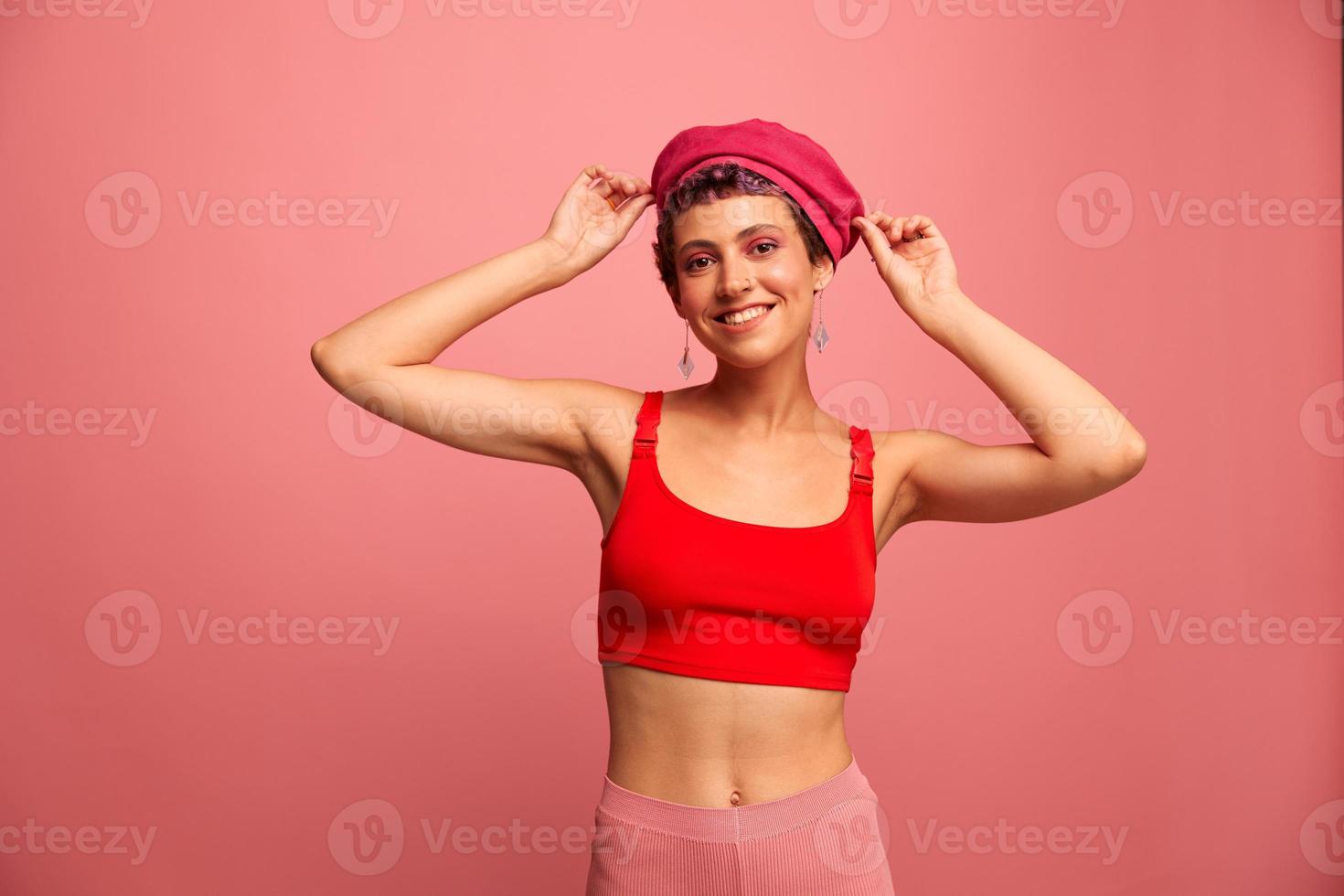 ung atletisk kvinna med en kort frisyr och lila hår i en röd topp och en rosa hatt med ett atletisk figur ler och grimaser som visar tecken med henne händer ser på de kamera på en rosa bakgrund foto