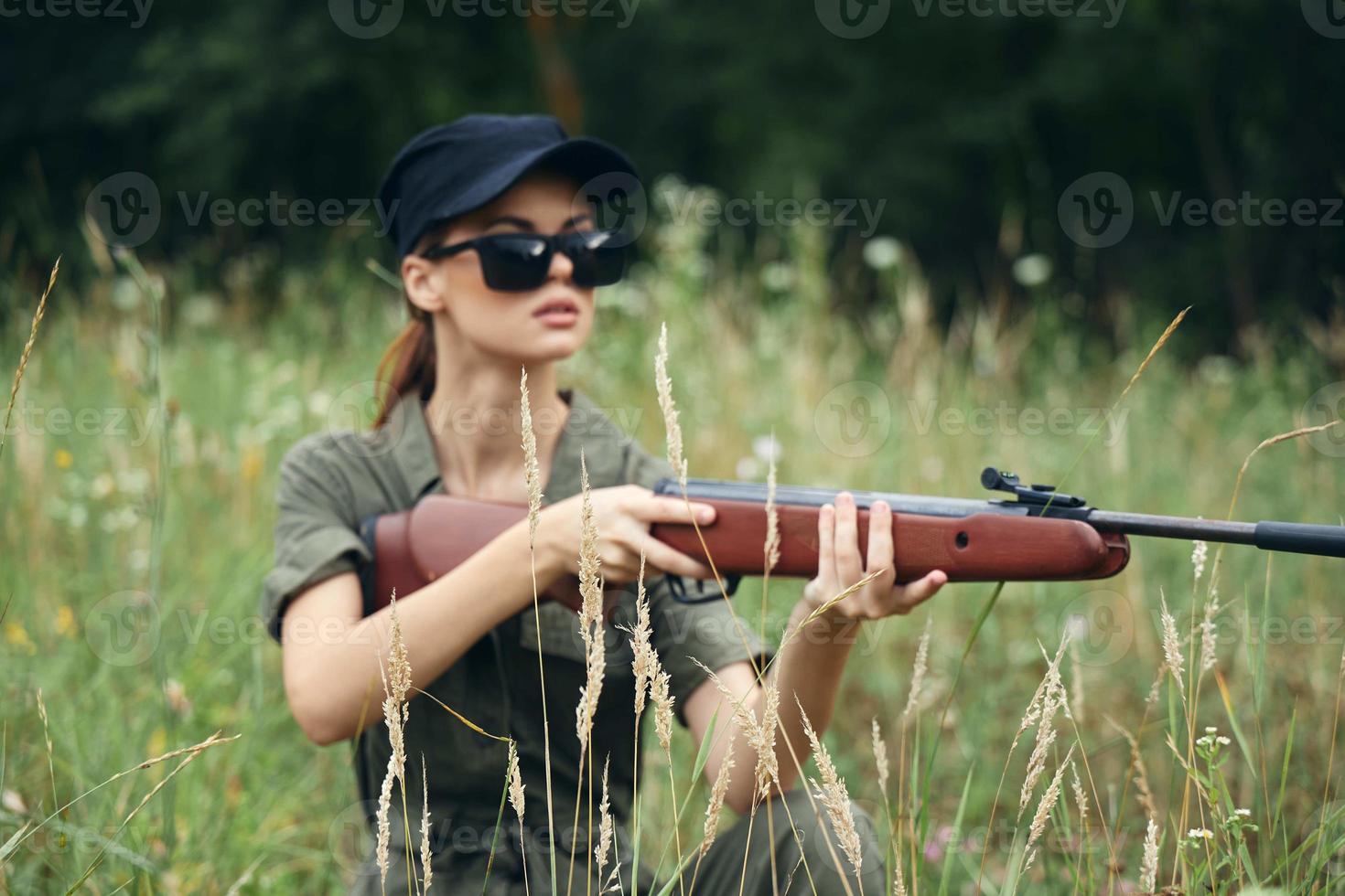 kvinna på utomhus- i mörk glasögon med vapen skydd jakt grön löv grön träd foto