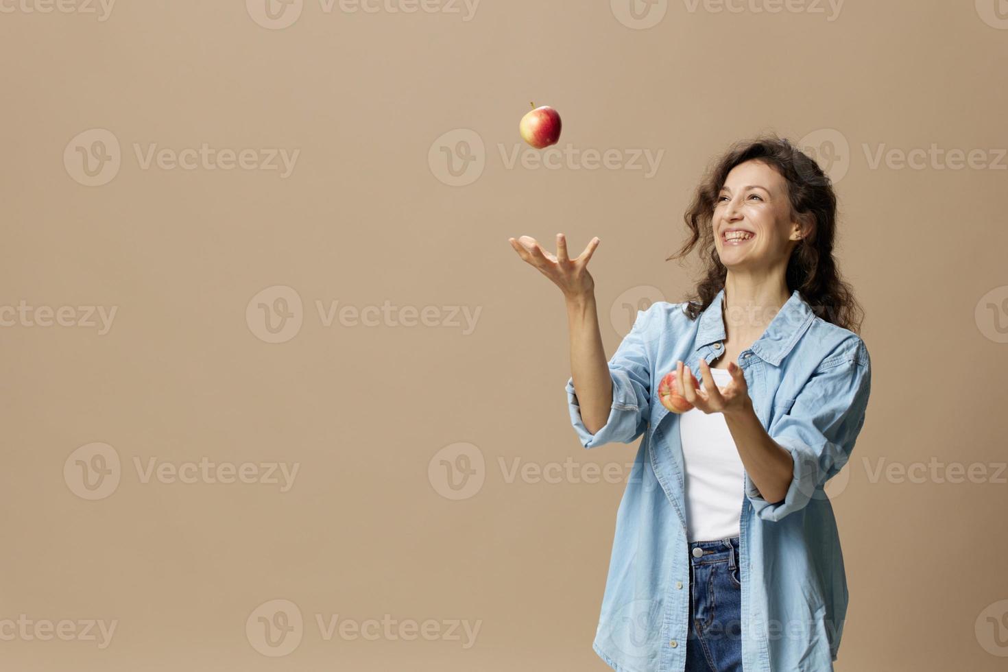 Lycklig leende fokuserade lockigt skön kvinna i jeans tillfällig skjorta jonglering med äpple Framställ isolerat på över beige pastell bakgrund. friska mat. naturlig miljövänlig Produkter begrepp. kopia Plats foto