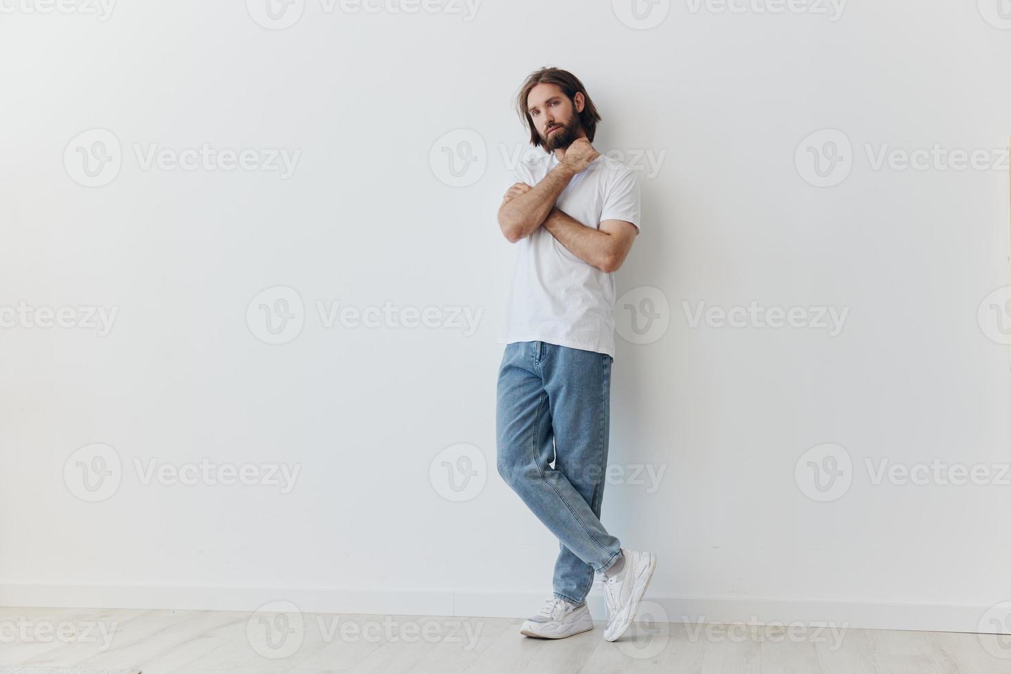 en man i en vit t-shirt och blå jeans står mot en vit vägg och poser foto