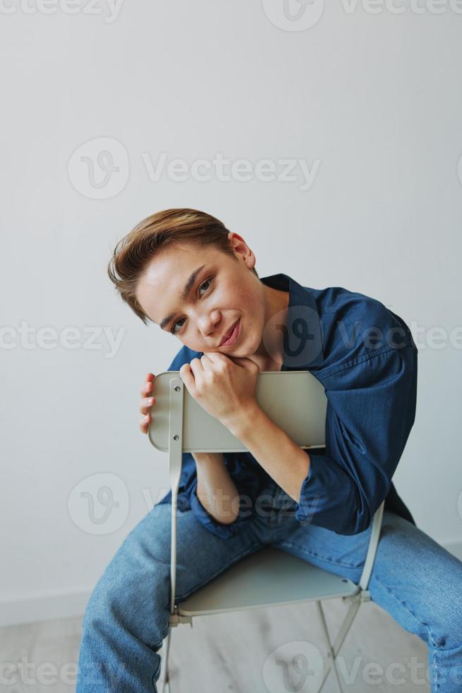 en ung kvinna Sammanträde i en stol på Hem leende med tänder med en kort frisyr i jeans och en denim skjorta på en vit bakgrund. flicka naturlig poser med Nej filter foto