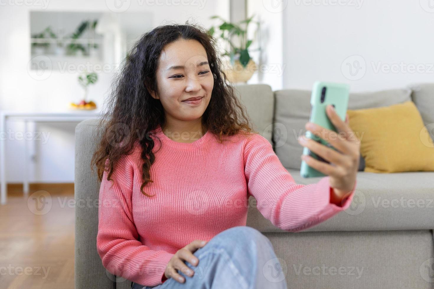 attraktiv leende asiatisk kvinna använder sig av smart telefon medan Sammanträde på de soffa på Hem. kommunikation och video ring upp begrepp. foto