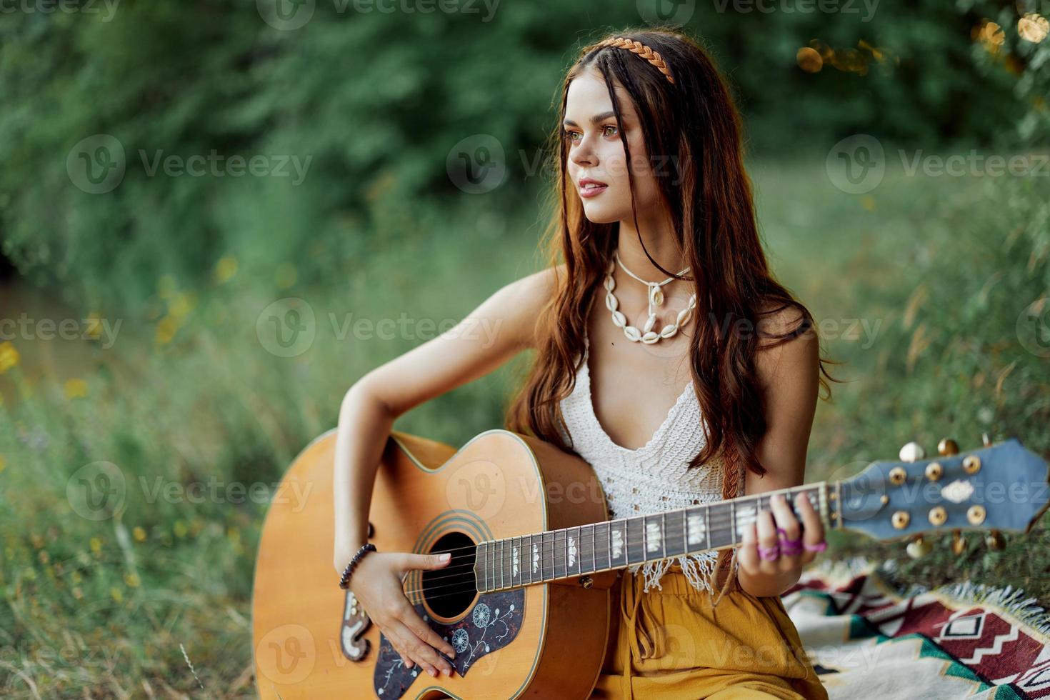 hippie kvinna leende och kramas henne gitarr i natur i de parkera i de solnedgång ljus i eko-kläder foto