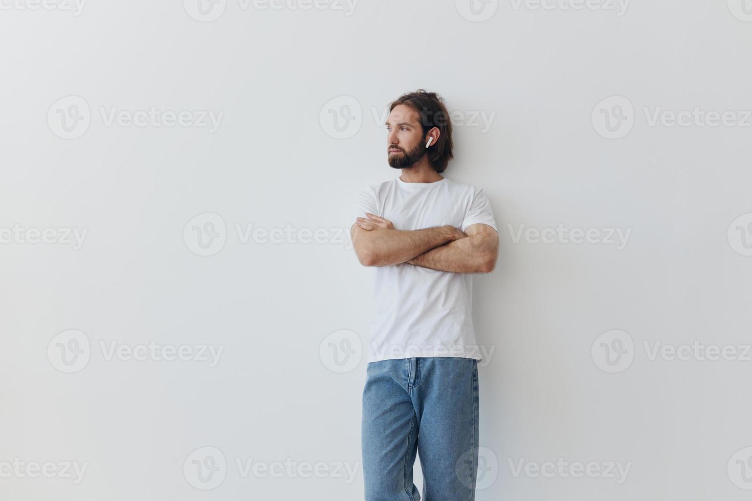 en man med en skägg och lång hår i en vit t-shirt och blå jeans står mot en vit vägg, lutande mot den och lyssnande till musik med trådlös vit hörlurar, stirrande eftertänksamt foto