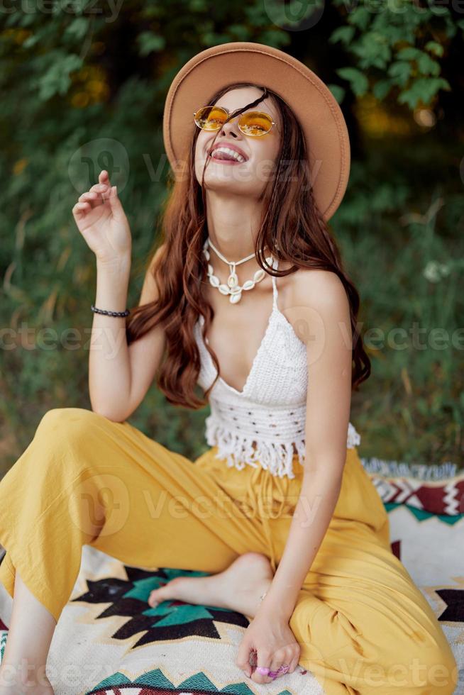 hippie kvinna i eco Kläder skrattar och vickar som hon sitter på en pläd utanför i de falla med en hatt och gul solglasögon foto