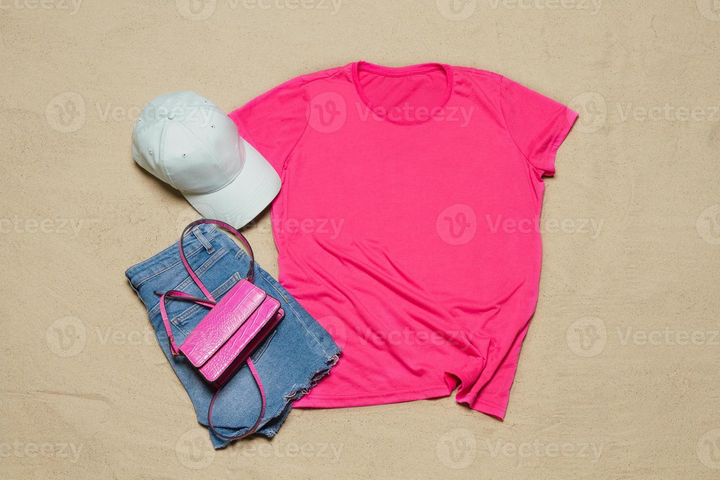 sand strand textur bakgrund. attrapp rosa sommar t-shirt utrusta kopia Plats. tom mall kvinna skjorta topp se. sommartid Tillbehör hatt, jeans. platt-lay närbild tshirt på stranden. Strand tid foto
