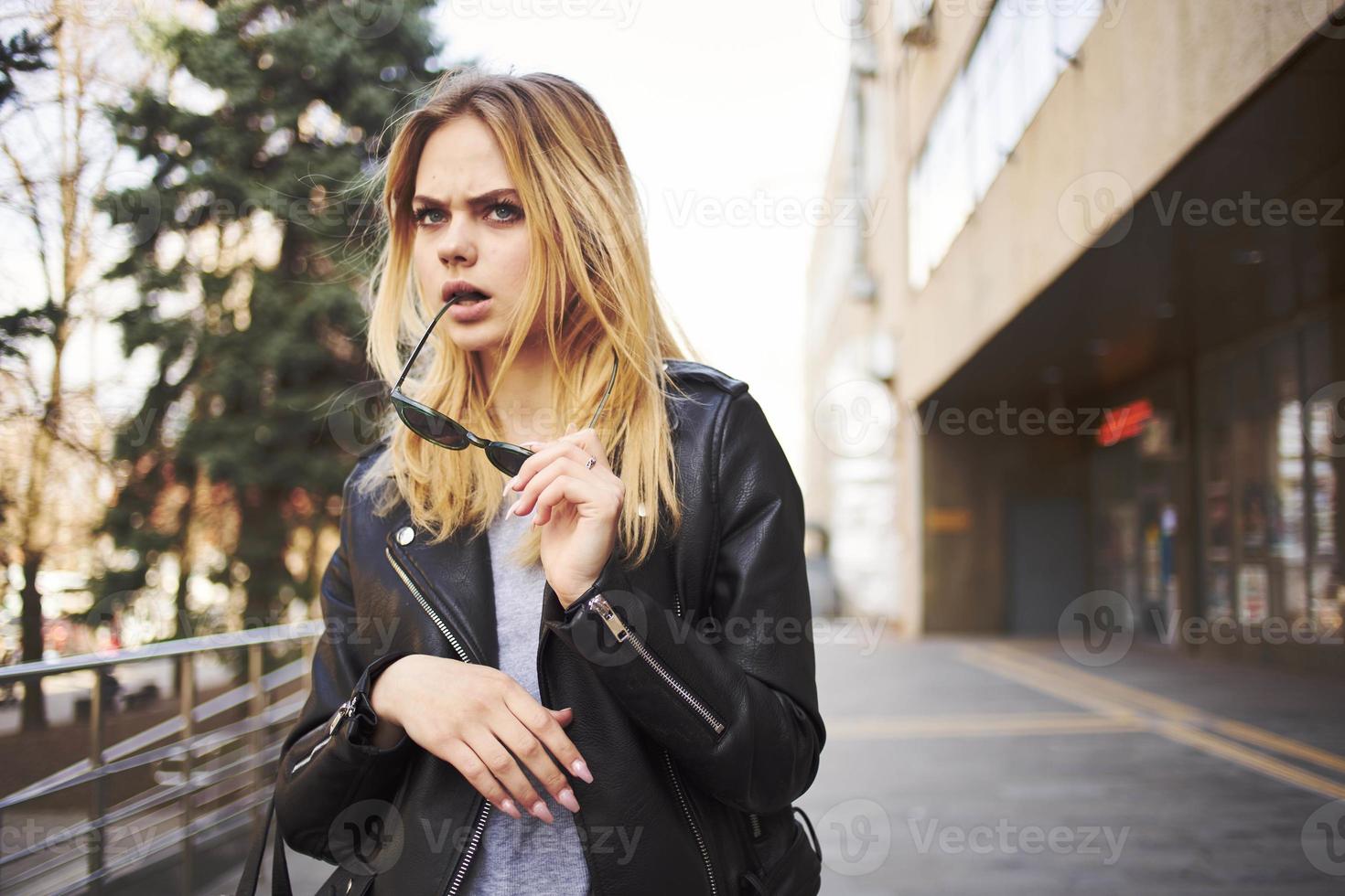 en blond kvinna i en läder jacka är gående på de gata i de stad nära de byggnad foto