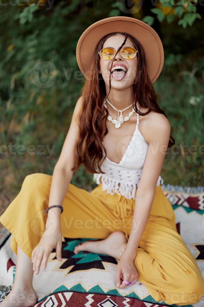 hippie kvinna i eco Kläder skrattar och vickar som hon sitter på en pläd utanför i de falla med en hatt och gul solglasögon foto