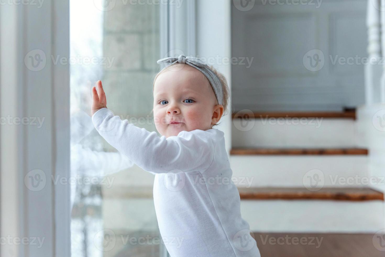liten krypande bebis flicka ett år gammal lokaliserings på golv i ljus ljus levande rum nära fönster leende och skrattande foto