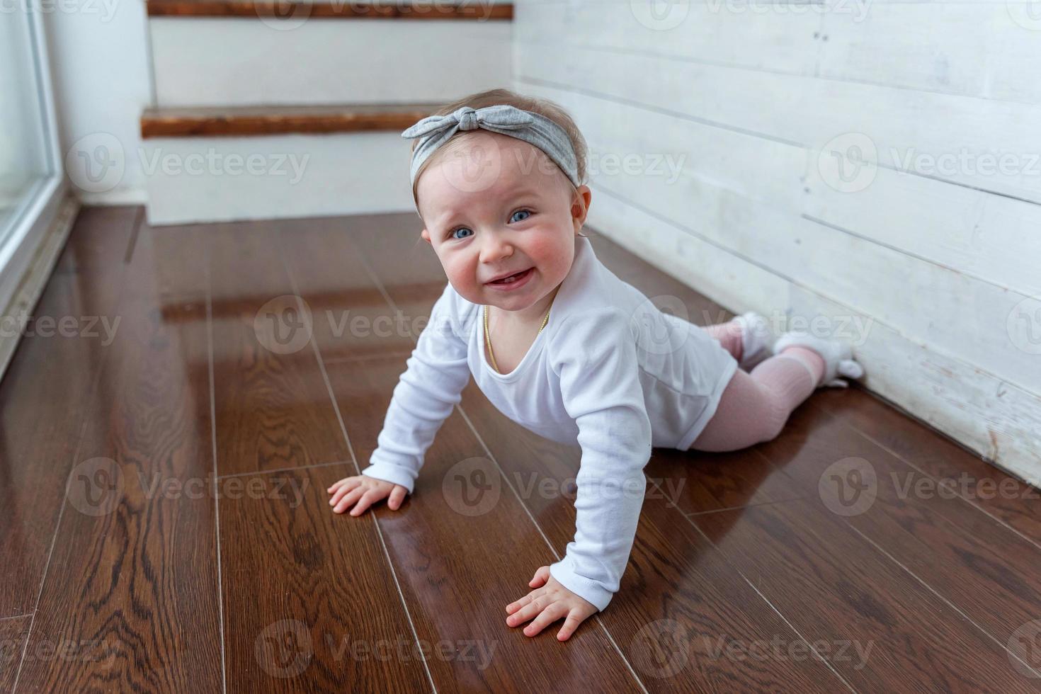 liten krypande bebis flicka ett år gammal lokaliserings på golv i ljus ljus levande rum nära fönster leende och skrattande foto