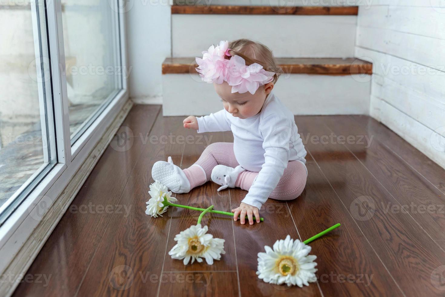 liten bebis flicka bär vår krans lokaliserings på golv i ljus ljus levande rum nära fönster och spelar med gerbera blommor foto
