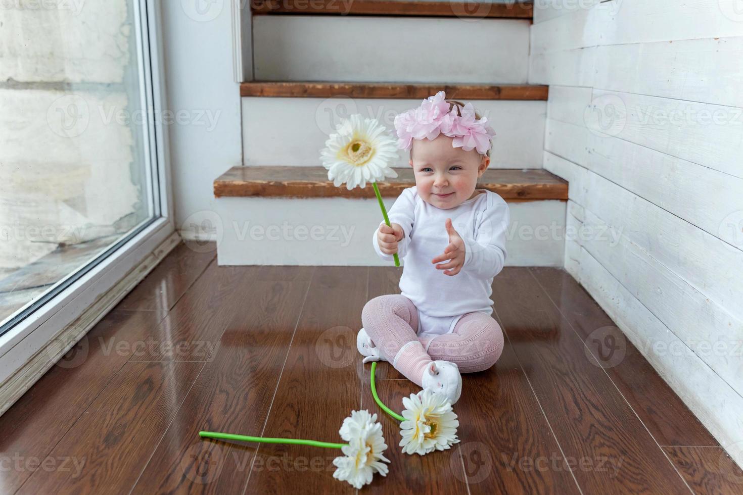 liten bebis flicka bär vår krans lokaliserings på golv i ljus ljus levande rum nära fönster och spelar med gerbera blommor foto