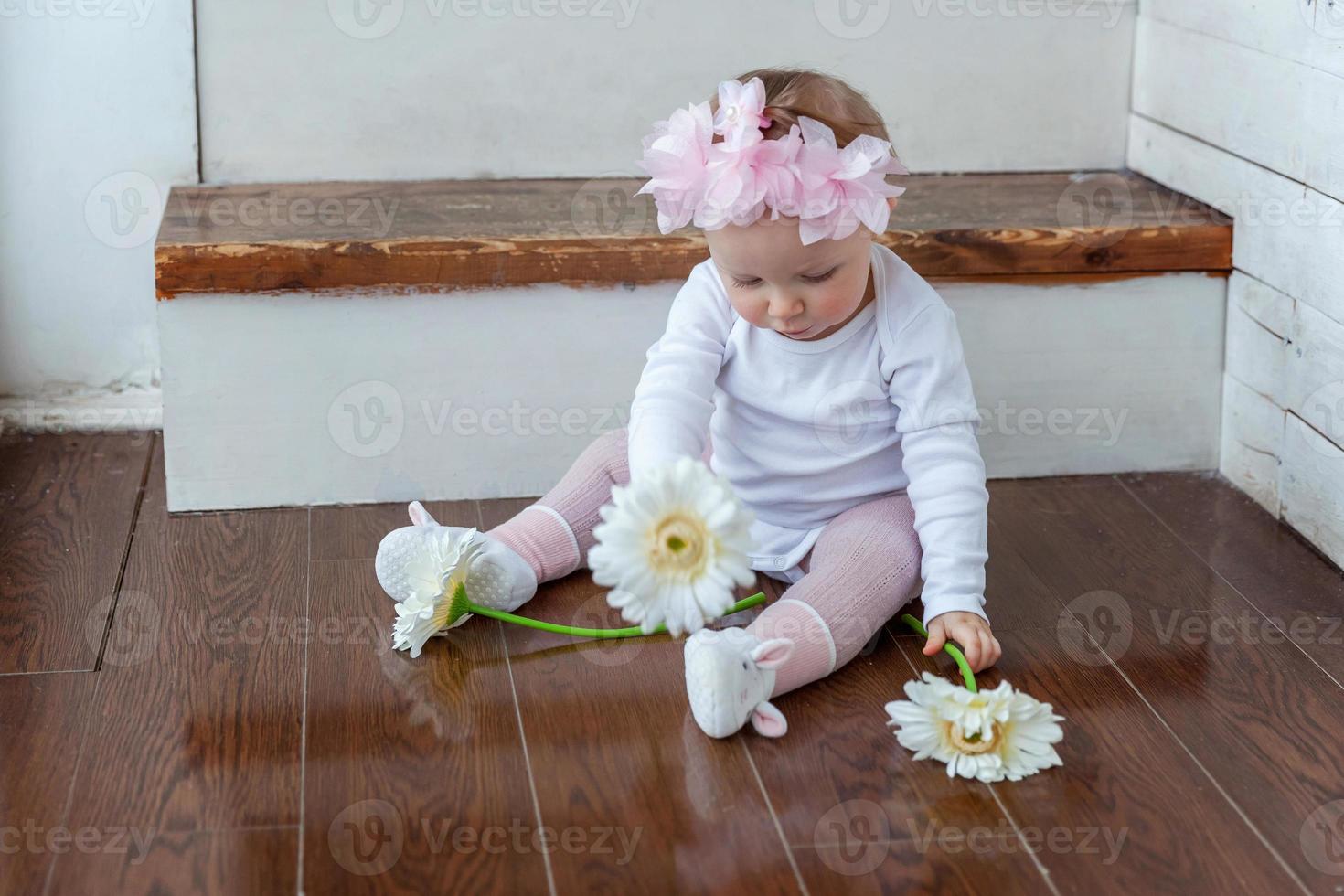 liten bebis flicka bär vår krans lokaliserings på golv i ljus ljus levande rum nära fönster och spelar med gerbera blommor foto