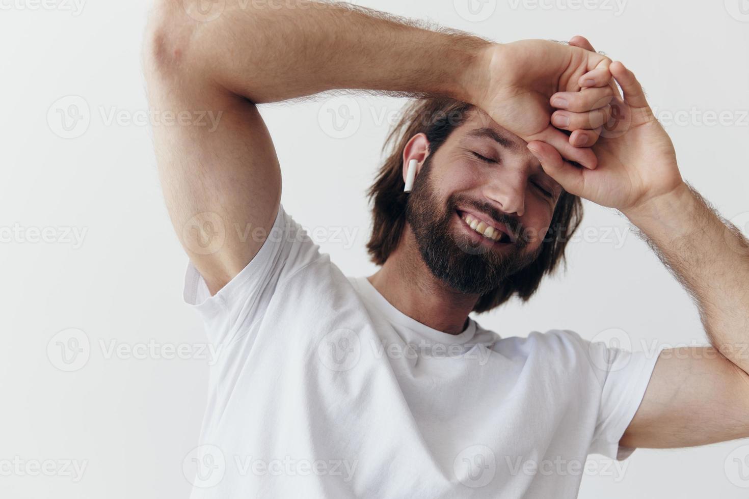 eleganta man i en vit t-shirt med trådlös hörlurar i hans öron har roligt lyssnande till musik leende på en vit bakgrund livsstil foto